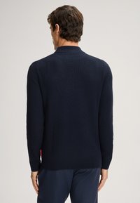 Dunkelblauer gestrickter Pullover mit Rippstruktur, langen Ärmeln, hohem Kragen und einem kleinen roten Etikett an der Seite.