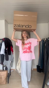 Caixa de cartão com o logótipo da Zalando, segurada acima da cabeça. Pessoa veste uma t-shirt gráfica cor-de-rosa e calças largas cinzentas numa divisão desarrumada com roupa.