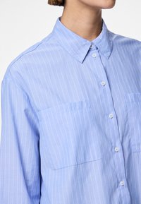 Vaaleansininen nappipaita, joka on valmistettu sileästä kankaasta ja jossa on pystysuorat valkoiset raidat, klassinen kaulus ja kaksi etutaskua.