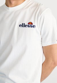 Camiseta de algodón blanca con cuello redondo y mangas cortas. Presenta un logotipo bordado de "ellesse" en color navy y un acento colorido.