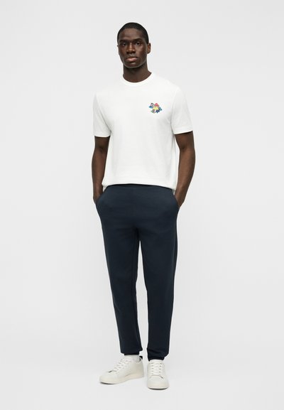 T-shirt blanc à manches courtes comportant un graphisme coloré sur la poitrine gauche, associé à un pantalon de jogging bleu marine foncé et des baskets blanches.
