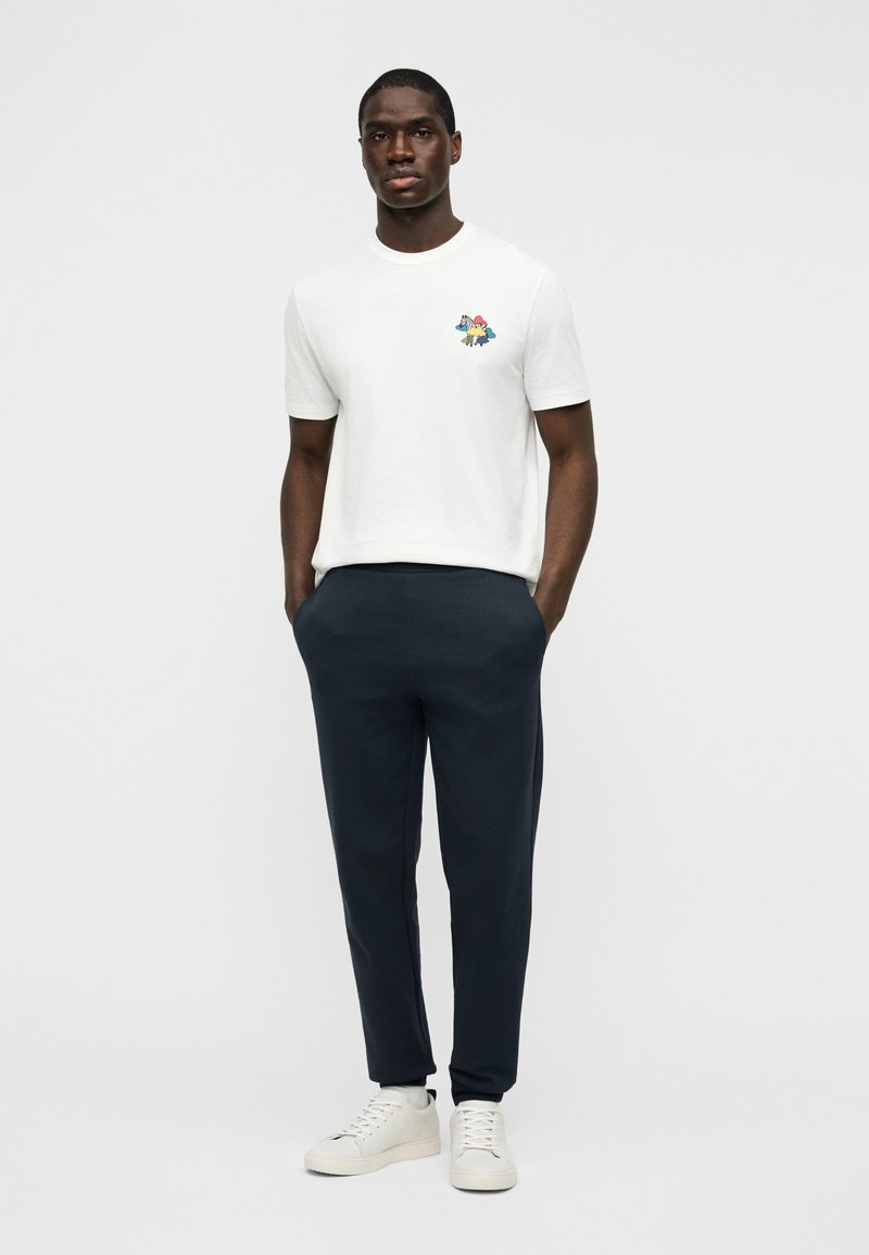 Λευκό κοντομάνικο μπλουζάκι με πολύχρωμο σχέδιο στο αριστερό στήθος, συνδυασμένο με σκουρόχρωμο μπλε παντελόνι jogger και λευκά αθλητικά παπούτσια.