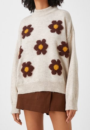 Pull oversized beige clair avec un col haut, orné de motifs floraux marron foncé et jaunes, et doté de poignets et d'un ourlet côtelés. Texture douce.