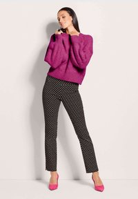 Fuchsia gestrickter Pullover mit Puffärmeln, kombiniert mit schwarzen Hosen mit Punkten und leuchtend pinken High Heels. Glatte Textur, taillierte Passform.