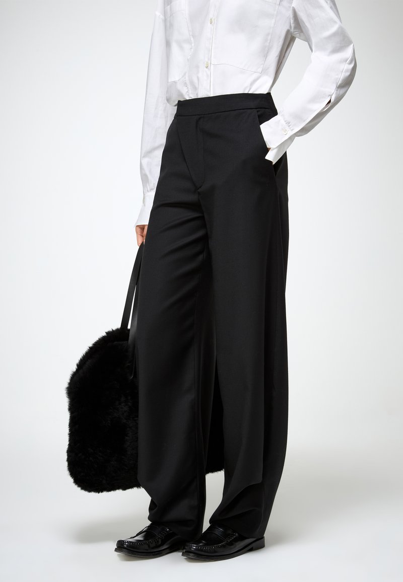 Pantalons larges noirs avec une finition en tissu lisse, associés à une chemise blanche à boutons et un sac noir duveteux. Les chaussures sont noires et polies.