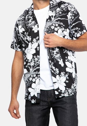 Korte mouw zwarte shirt met witte bloemenpatroon. Katoenen materiaal, relaxte pasvorm en een knoopsluiting kraag. Gedragen over een wit shirt.