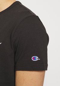 Musta puuvillainen t-paitaja, jossa on lyhyet hihat ja pieni sininen ja punainen brodeerattu Champion-logo vasemmassa hihassa.