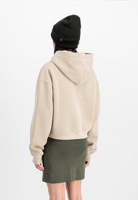 Beige īsi izgriezta kapuci ar kapuci un platām piedurknēm, saskaņota ar tumši zaļu pieguļošo svārku. Melna beanie papildina izskatu.