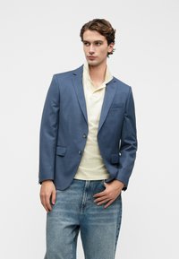 Μπλε σακάκι με κανονικούς γιακάδες, δύο κουμπιά και μπροστινές τσέπες, γεμάτο από μια ανοιχτό κίτρινη πόλο, συνδυασμένο με ανοιχτό μπλε τζιν.