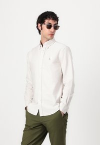 Camisa de botones blanca a rayas, de tela ligera, con un pequeño logo en el pecho. Combinada con pantalones verde oliva y gafas de sol.