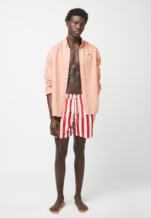 Chemise longue à manches longues rose clair avec bouton à l'avant, portée sur un short de bain rouge et blanc à rayures avec cordon de serrage. Le modèle est pieds nus.