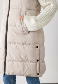 Beige puffer vest med frontglidelås, sidelommer og tonede ribbestrikkede ermer. Utdifferentierte svarte trykknapper på sidesømmene. Overlagt på denim.