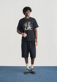 Μαύρο βαμβακερό t-shirt με γραφικό τύπωμα και λογότυπο, συνδυασμένο με μαύρο σορτς μέχρι το γόνατο. Μαύρα αθλητικά παπούτσια και μπεζ κάλτσες ολοκληρώνουν το σύνολο.