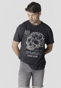 Man wearing black T-shirt with geometric skull design and text, paired with gray rolled-up shorts, looking down with hands behind back.