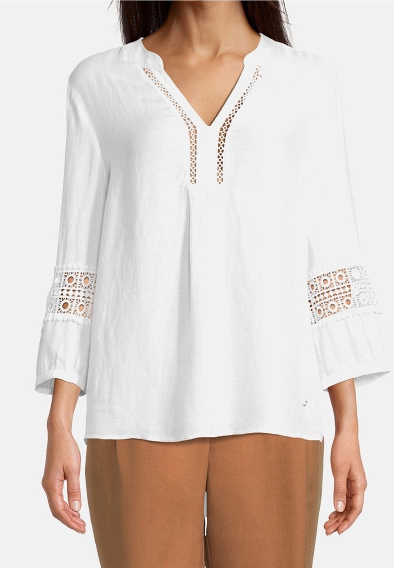 Blusa bianca in lino con scollo a V, caratterizzata da dettagli in pizzo lungo il collo e maniche a campana 3/4 con intricati motivi ritagliati.