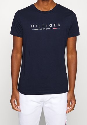 Person trägt ein dunkelblaues T-Shirt mit dem Text "HILFIGER NEW YORK" und farbigen Balken, kombiniert mit weißen Hosen, die einen teilweisen Logotext zeigen.