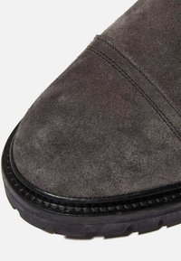 Bottine en suède gris avec un bout rond et une semelle en caoutchouc noire contrastante. Présente des coutures visibles sur la partie supérieure et une texture lisse.