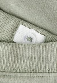 Jasnozielona tkanina o miękkiej fakturze. Posiada biały tag z guzikiem, z subtelnym prążkowanym wzorem wzdłuż krawędzi.