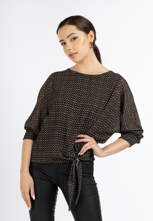 Woman with dark hair in a low ponytail wearing a black patterned blouse with a front tie and black pants, posing with one hand on hip.
