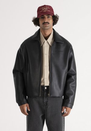 Mand med krøllet hår og overskæg iført en vinrød kasket, beige skjorte med knapper, sort læderjakke og mørke jeans, stående foran en hvid baggrund.