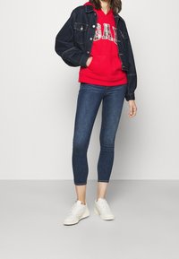 Červená mikina s kapucí s bílým nápisem "GAP", tmavá džínová bunda se dvěma předními kapsami, úzké modré džíny a bílé tenisky.