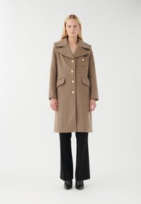 Manteau en mélange de laine marron avec un large col, six grands boutons, poches latérales et une silhouette droite, associé à un pantalon noir évasé.