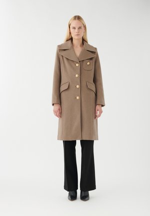 Beige Wollmantel mit breitem Kragen, großen goldenen Knöpfen und zwei vorderen Taschen. Kombiniert mit schwarzten Schlaghosen und spitzen schwarzen Schuhen.