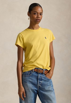 Femeie purtând un tricou galben cu mâneci scurte și un logo mic, blugi albaștri, o curea maro țesută și o singură cercei picătură, pozând pe un fundal simplu.