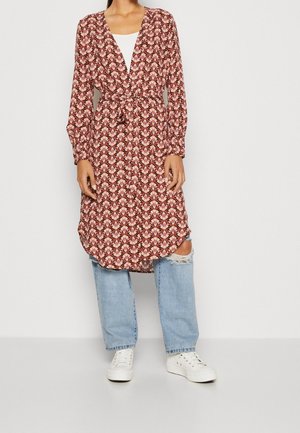 Personne portant une robe portefeuille à manches longues à motifs par-dessus un haut blanc et un jean bleu clair déchiré avec des baskets blanches sur un fond uni.