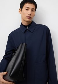 Granatowa zewnętrzna kurtka z kołnierzykiem, zapinana na guziki z przodu i z bocznymi kieszeniami, w połączeniu z gładką czarną torebką noszoną w prawej ręce.