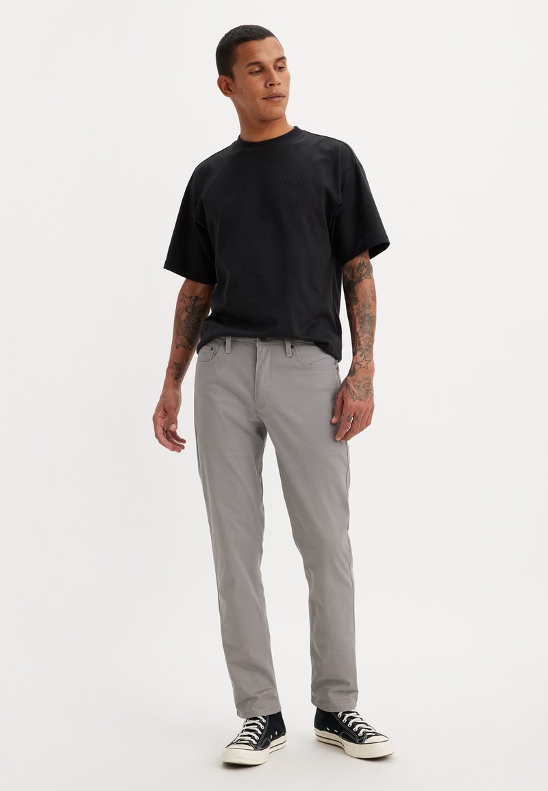 T-shirt noir à manches courtes, pantalon slim gris clair, et baskets noires avec semelles blanches. Présente des tatouages sur les bras.