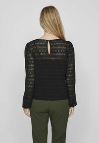 Mustmustrek pikkade varrukatega ülaosa, millel on avatud seljaosa detail ja tekstuurilised lillemustrid, koos tumeroheliste pükstega.