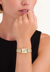 Reloj de pulsera rectangular dorado con una cara suave y de color claro y una correa de eslabones de metal, descansando sobre una mano cerca de la barbilla.