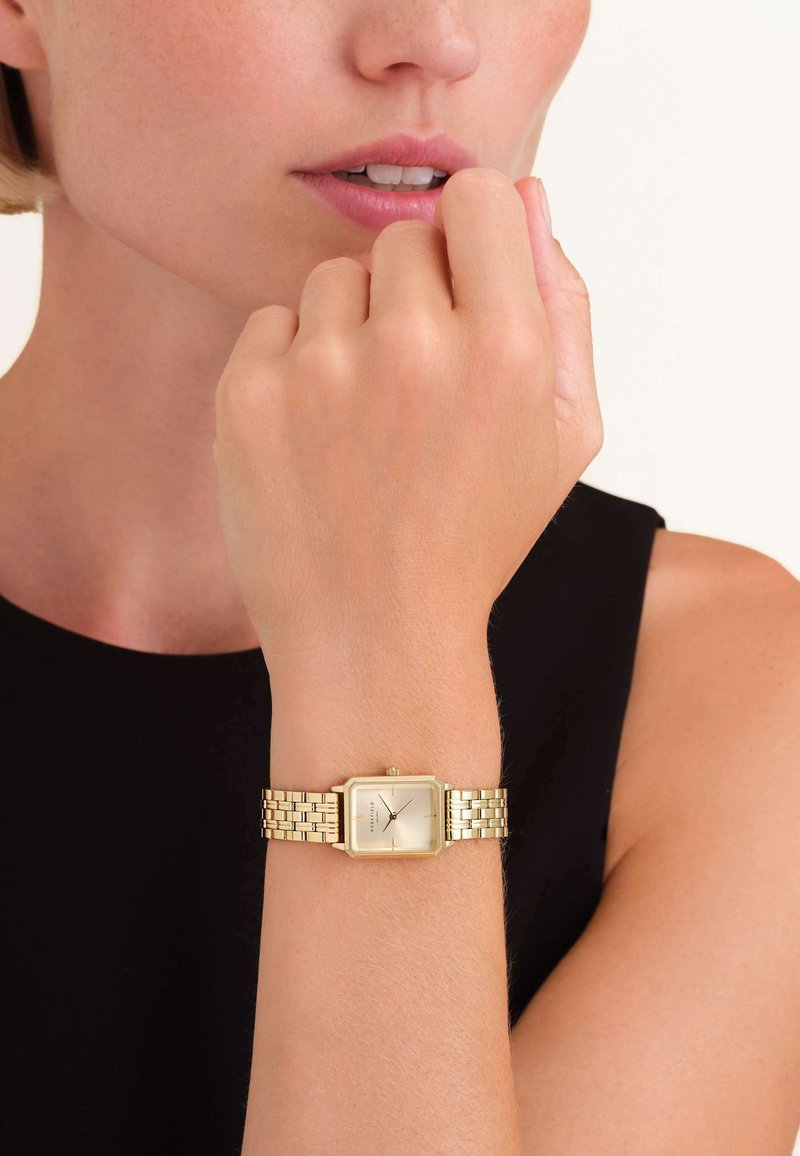 Reloj de pulsera rectangular dorado con una cara suave y de color claro y una correa de eslabones de metal, descansando sobre una mano cerca de la barbilla.