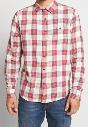 Man in een rood, wit en roze geruite overhemd met een borstzak en blauwe jeans, staand met de armen ontspannen langs de zij.