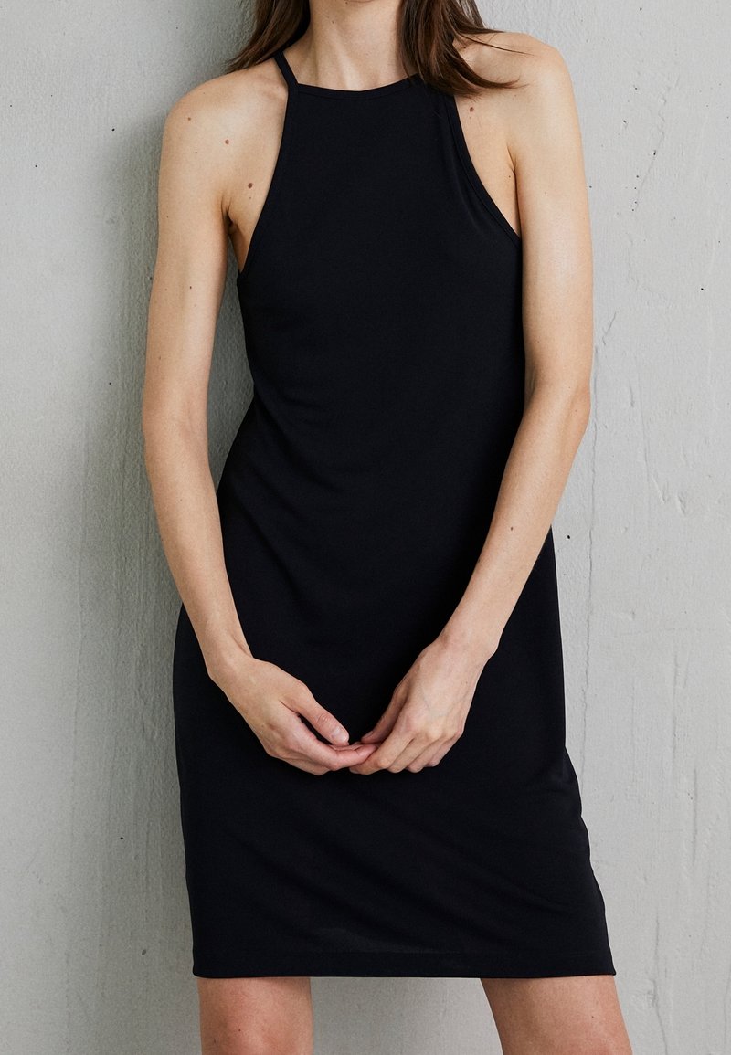 Jeune femme portant une robe noire sans manches à col halter, debout contre un mur gris clair texturé, les mains jointes devant.