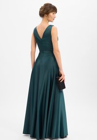 Lange mouwloze jurk in donker teal satijn met een V-rug ontwerp. De vloeiende rok valt tot op de vloer, aangevuld met een zwarte clutch.