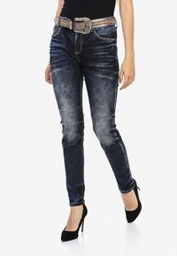 Jean en denim bleu foncé avec une coupe slim, texture délavée, poches accentuées et une ceinture décorative en argent. Porté avec des chaussures à talons hauts noires.