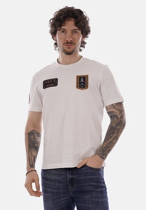 Uomo con capelli ricci che indossa una maglietta bianca con due spille e tatuaggi su entrambi gli avambracci, vestito con jeans blu, con una mano in tasca.