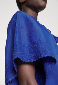 Garbò blu ricamato con dettagliati motivi a foglia sull'orlo, morbida texture e design fluido. Il tessuto si adagia intorno alla spalla.