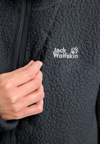 Schwarze Fleecejacke mit strukturierter Oberfläche, ausgestattet mit einem Reißverschluss und einem silbernen "Jack Wolfskin"-Logo auf der Brust. Nahaufnahme einer Hand, die den Reißverschluss anpasst.