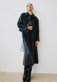 Czarna skórzana krótka kurtka z kołnierzem, zestawiona z pasującą długą spódnicą i wysokimi kozakiem do kolan. Kopertówka posiada długie frędzle jako ozdobę.