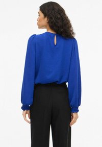 Blusa blu con texture, maniche a sbuffo, apertura a goccia sul retro e polsini elastici, abbinata a pantaloni neri a vita alta.