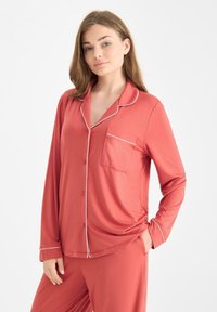 Een vrouw in een koraalrode pyjamastel met witte biesdetails, staand met één hand in de zak tegen een effen lichte achtergrond.
