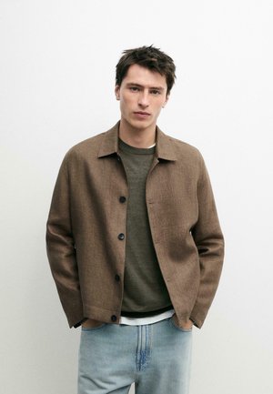 Jeune homme aux cheveux courts et foncés portant une veste marron boutonnée sur un pull vert olive et un jean bleu clair, debout les mains dans les poches.