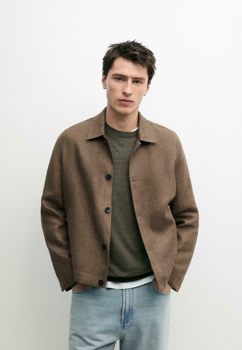 Jeune homme aux cheveux courts et foncés portant une veste marron boutonnée sur un pull vert olive et un jean bleu clair, debout les mains dans les poches.