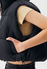 Osoba v černé péřové vestě přes béžovou košili s krátkým rukávem, s rukou v kapse vesty, na bílém pozadí.