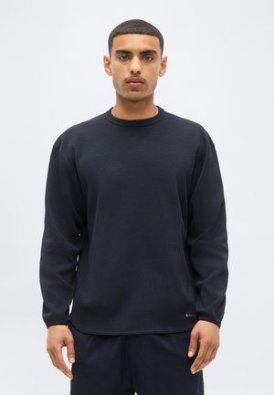 Jeune homme aux cheveux courts portant un t-shirt noir uni à manches longues en tissu gaufré, debout sur un fond gris clair.