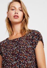 Noor naine heledate juustega kannab suuri kuldseid rõngaskõrvarõngaid ja musta lühikeste varrukatega lillelist pluusi oranžide, valgete ja punaste lilledega.