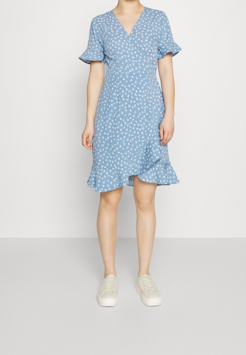 Femme portant une robe portefeuille bleue à fleurs longueur genou avec des manches courtes volantées et des baskets blanches, debout devant un fond blanc.